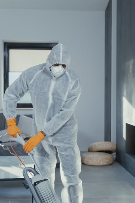 A professional cleaner dressed in a white protective suit, gloves, and shoe covers uses a wet vacuum or carpet extractor to deep clean a beige carpet in a bright, modern living room. The room features