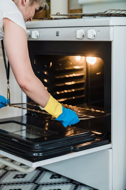 A person wearing a white top and yellow and blue rubber gloves is cleaning the inside of a white oven with a wire rack on a kitchen floor featuring a black and white patterned tile. The oven's interio